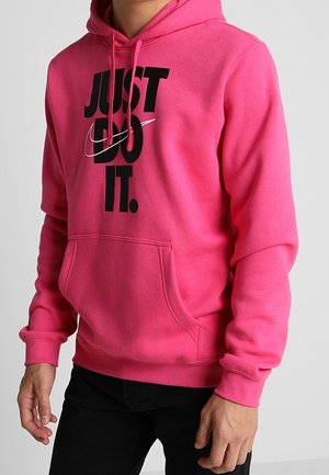 Osoba nosząca jasnoróżową bluzę z kapturem z czarnym napisem „JUST DO IT.” i białym logo Nike na przedniej stronie, w połączeniu z czarnymi spodniami.