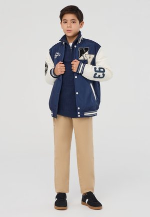 Giacca varsity blu navy e crema con polsini a coste, scritte ricamate e un design sportivo abbinata a pantaloni beige e sneakers nere.