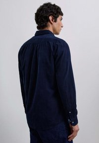 Camisa de pana azul marino con yugo trasero, puños abotonados y dobladillo recto, que presenta una textura suave y un brillo sutil.