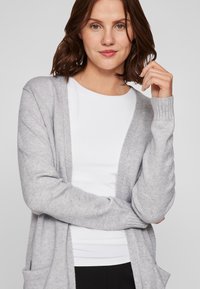 Hallide hall kardigan, millel on lõdvestunud sobivus, avatud esiosa, sooniku mansetid ja kaks esist taskut, valge särgi peal. Ühtlane tekstuur.