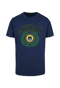 Tamsiai mėlyna medvilninė marškinėliai su užrašu „MICHIGAN“ ryškiai geltonomis raidėmis virš apvalaus emblemos su laurų lapais ir universiteto antspaudu.