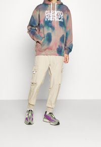 Mikina s kapucí a tie-dye vzorem v růžových, modrých a zelených tónech s bílým logem; béžové cargo kalhoty; šedé a fialové tenisky se zelenými akcenty.