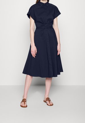 Mujer con un vestido azul marino hasta la rodilla, de mangas cortas y con detalle de cruzado, combinado con sandalias planas marrones de punta abierta.