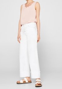 Kvinna iklädd en ärmlös ljusrosa topp med scallop-dekoration, högmidjade vida vita jeans och vita sandaler med remmar.