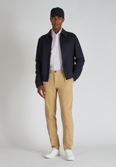 Joven vestido con una chaqueta azul marino, camisa de botones a rayas, pantalones beiges, zapatillas blancas y una gorra azul marino, de pie contra un fondo liso.
