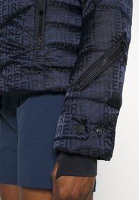 Námořnická prošívaná bunda s výrazným vzorem "OHEŇ", kapsou na zip na manžetě a černými patentními knoflíky jako ozdobou. Hladká nylonová textura, prošívaný design.