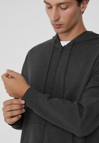 Sudadera con cremallera de color gris oscuro, fabricada con un tejido texturizado, con un bolsillo tipo canguro y cordones ajustables en la capucha. El modelo ajusta el puño.