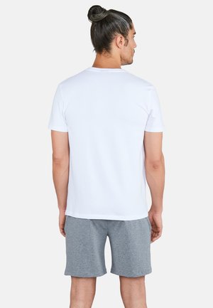 Mies, jolla on nutturakampaus, pukeutuneena yksinkertaiseen valkoiseen lyhythihaiseen t-paitaan ja harmaisiin shortseihin, seisoo selin valkoista taustaa vasten.