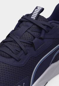Σκούρο μπλε αθλητικό παπούτσι Puma με ύφασμα από δίχτυ, χοντρές κορδόνια, λογότυπο στη γλώσσα και λευκή σόλα με μπλε λεπτομέρειες, εμφανίζεται σε κοντινό πλάνο.