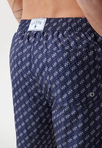 Pantaloni da bagno blu navy con motivo geometrico bianco. Vita elasticizzata con etichetta "GUESS", tasca posteriore e dettaglio con occhiello in metallo.