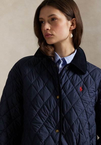 Femeie cu păr castaniu până la umeri, purtând o jachetă bleumarin matlasată peste o cămașă bleu deschis cu guler, cu o cerceată albă atârnândă.