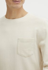 Chemise en tricot beige à manches longues avec un col rond et une poche en tissu texturé sur la poitrine gauche. Tissu lisse avec des détails de couture subtils.