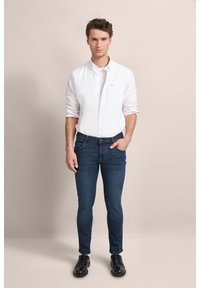 Biała koszula zapinana na guziki, niebieskie dżinsy i czarne eleganckie buty. Model stoi na neutralnym tle, prezentując dopasowany i swobodny strój.