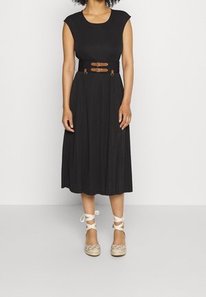 Woman wearing a black midi dress with cap sleeves and a wide belt with two brown buckles, paired with lace-up espadrille flats.