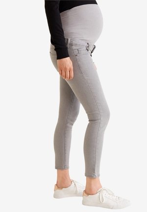 Schwangere Person trägt graue Umstandsjeans und weiße Sneaker, steht seitlich mit der Hand am Körper.