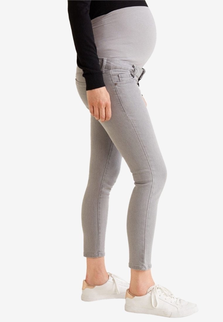 Personne enceinte portant un jean de maternité gris et des baskets blanches, debout de profil avec la main reposant le long du corps.