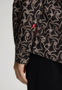 Camicetta in tessuto nero con un motivo a corda beige, dotata di polsino con bottone e un'etichetta rossa vicino alla cucitura. Texture liscia, vestibilità sagomata.