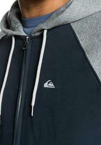 Navy zip-up hoodie with gray sleeves and drawstrings. Features a textured fabric with a small logo on the chest. Front zipper closure.