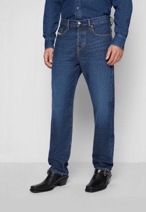 Mężczyzna w ciemnoniebieskich jeansach denim, pasującej koszuli denim i czarnych skórzanych botkach z klamrami, stojący na szarej podłodze.