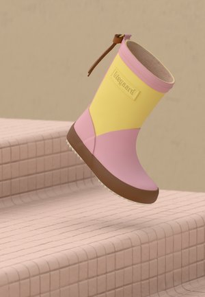 Botte de pluie rose et jaune pour enfant avec semelle marron, montrée en l'air au-dessus d'un escalier carrelé rose.