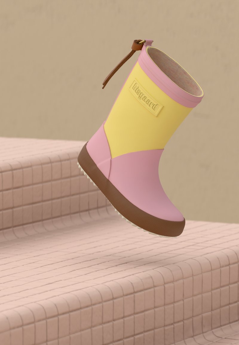 Botte de pluie rose et jaune pour enfant avec semelle marron, montrée en l'air au-dessus d'un escalier carrelé rose.