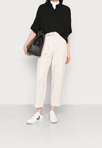 Camicia nera over size con bottoni, pantaloni crema a vita alta e sneakers bianche con accenti blu navy e rossi. Porta una borsa in pelle nera.