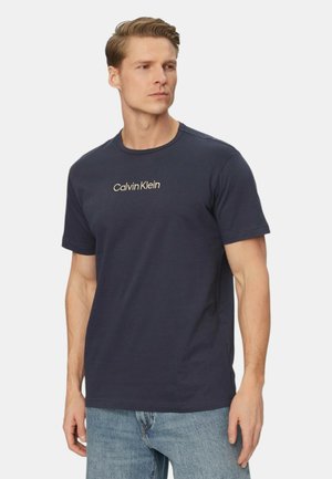 Uomo che indossa una t-shirt Calvin Klein blu navy e jeans blu, che guarda di lato su uno sfondo chiaro e uniforme.
