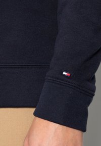 Fleece di colore navy con polsini a coste; piccolo emblema ricamato della bandiera nei colori rosso, bianco e blu sul polsino; dettagli di cuciture pulite.
