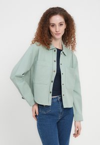 Veste vert menthe en coton, avec fermeture à boutons, deux poches avant et une coupe décontractée. Associée à un jean bleu.