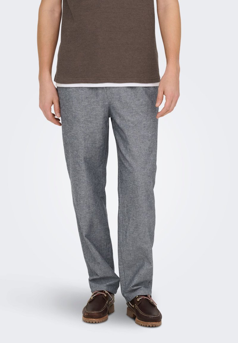 Homme portant un pantalon gris droit, des chaussures bateau marron foncé et une chemise marron à manches courtes, debout devant un fond uni.
