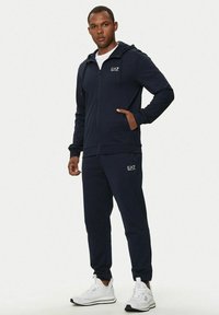 Felpa con zip blu navy e pantaloni jogger coordinati in tessuto morbido, con accenti del logo e tasche a scomparsa, abbinati a scarpe da ginnastica bianche.