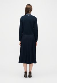 Γυναίκα που στέκεται πλάτη στην κάμερα, φορώντας μπλε μακρυμάνικη μπλούζα και πλισέ μίντι φούστα με καφέ παπούτσια με τακούνι.