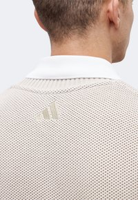 Πίσω μέρος του λαιμού και του άνω μέρους της πλάτης ενός ατόμου που φοράει ένα μπεζ υφασμάτινο πουλόβερ με λευκό γιακά και κεντημένο λογότυπο κοντά στη λαιμόκοψη.