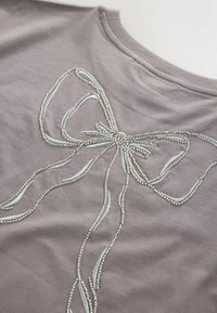 Top de tela gris con un gran lazo decorativo hecho de cuentas plateadas y bordado en azul claro, junto con detalles fluidos similares a cintas.