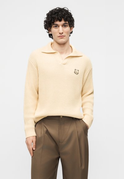 Jeune homme aux cheveux courts et bouclés portant un pull en maille crème avec un petit logo de loup et un pantalon plissé marron, les mains dans les poches.