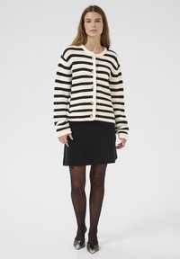 Zwart en crèmekleurig gestreepte cardigan met een ronde hals, grote knopen en oversized mouwen, gedragen over een zwarte rok en panty. Zilveren schoenen.