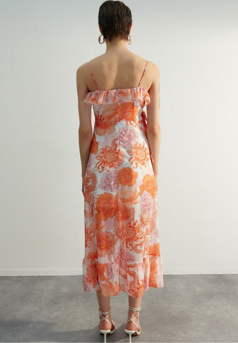 Femme aux cheveux courts portant une robe midi sans manches avec volants, orange et rose à motifs floraux, et des sandales blanches à talons, se tenant contre un mur clair uni.