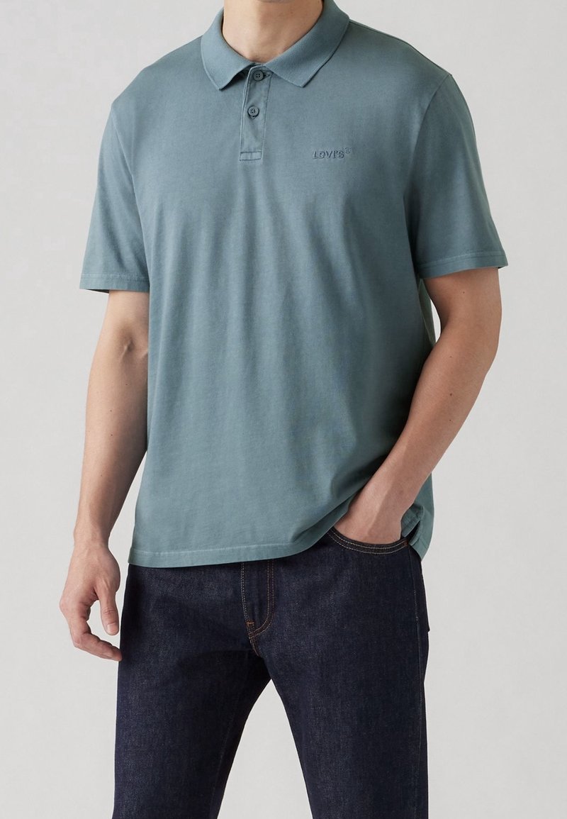 Homme portant un polo manches courtes de couleur sarcelle atténuée et un jean bleu foncé, une main dans la poche, fond neutre.