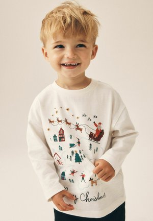 Smilende ung dreng med blondt hår iført en hvid langærmet julesweater med julemand, rensdyr, træer og festlige huse.