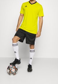 Camiseta atlética amarilla con cuello en V negro, pantalones cortos de malla negros, calcetines blancos con rayas negras y zapatos de fútbol negros, de pie junto a un balón de fútbol.