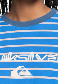 Blårandig t-shirt med rundad halsringning, med vit text och logotyp på bröstet. Tyget verkar slätt och lätt.