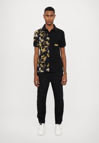 Svart polo skjorte med delt design: venstre side med gull- og hvit blomsterdesign, høyre side i solid svart. Modellen har på seg svarte bukser og hvite joggesko.