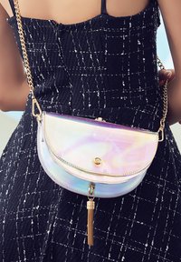 Iridescentna torbica u obliku polumjeseca s zlatnom lančićem, koja ima kružićasti zatvarač i ukras s resicama, pričvršćena na uzorčanu crnu odjeću.