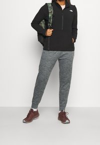 Person klädd i svart zip-up jacka, grå mjukisbyxor, maroon sneakers och bär en grön kamouflage ryggsäck på ena axeln.