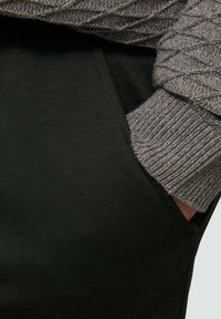 Dunkelgrüne Hose mit einer Seitentasche, kombiniert mit einem strukturierten grauen Strickpullover mit einem Rautenmuster. Nahaufnahme.