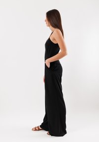 Tutina nera con spalline sottili, vestibilità ampia, tasche laterali e una texture liscia, abbinata a sandali marroni con cinturini.