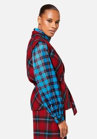 Rood en blauw geruit vest gelaagd over een bijpassende geblokte blouse; voorzien van knopen, een gebroken taille en poffende mouwen.
