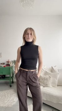 Črni brezrokavnik z visokim ovratnikom, kombiniran z rjavimi širokimi hlačami, ki imajo gladko teksturo in krojeno prileganje. Nevtralno ozadje z mehkimi pohištvenimi dodatki.