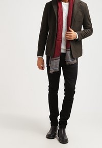 Braunes Sakko über weißem, kabelgestricktem Pullover, schwarze Jeans und schwarze Stiefel. Scharlachroter Schal mit blauen und grauen Akzenten um den Hals drapiert.