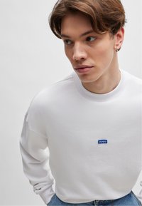 Sweatshirt blanc avec col rond côtelé, épaules tombantes, et logo "HUGO" bleu sur la poitrine ; porté avec un jean en denim bleu.